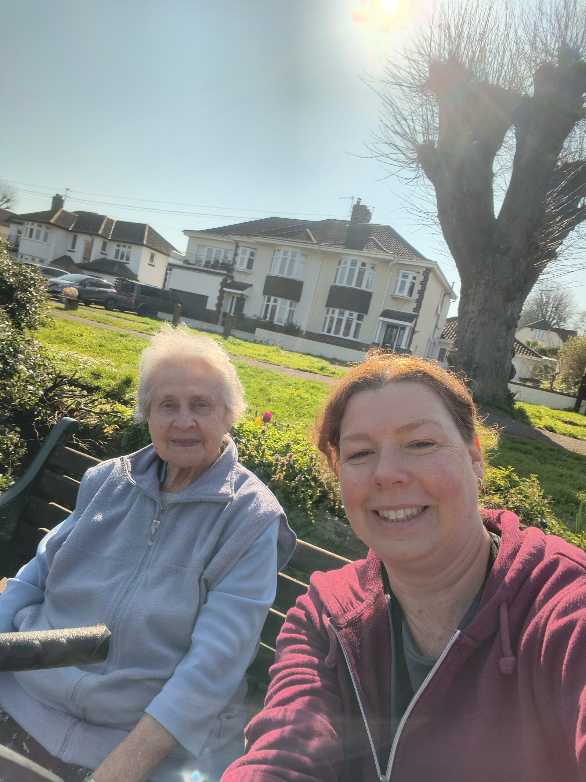 Resident and carer sitting on a bench in the park taking a selfie scaled