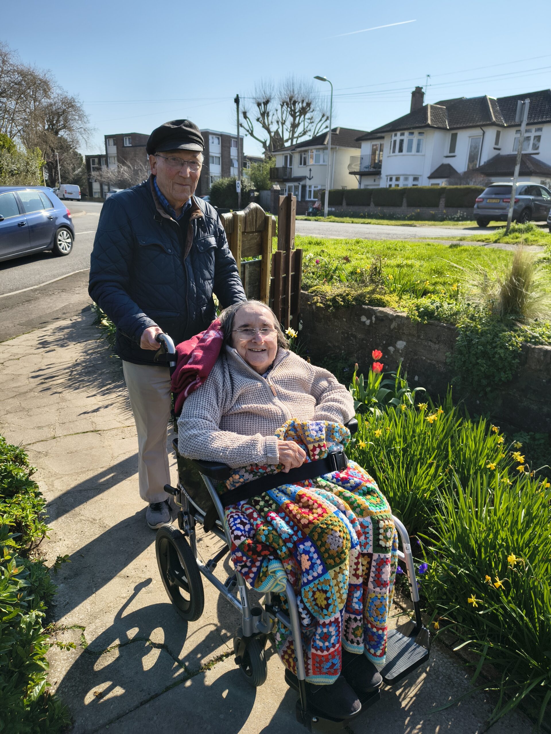 Residents one pushing a wheelchair with resident in a park scaled