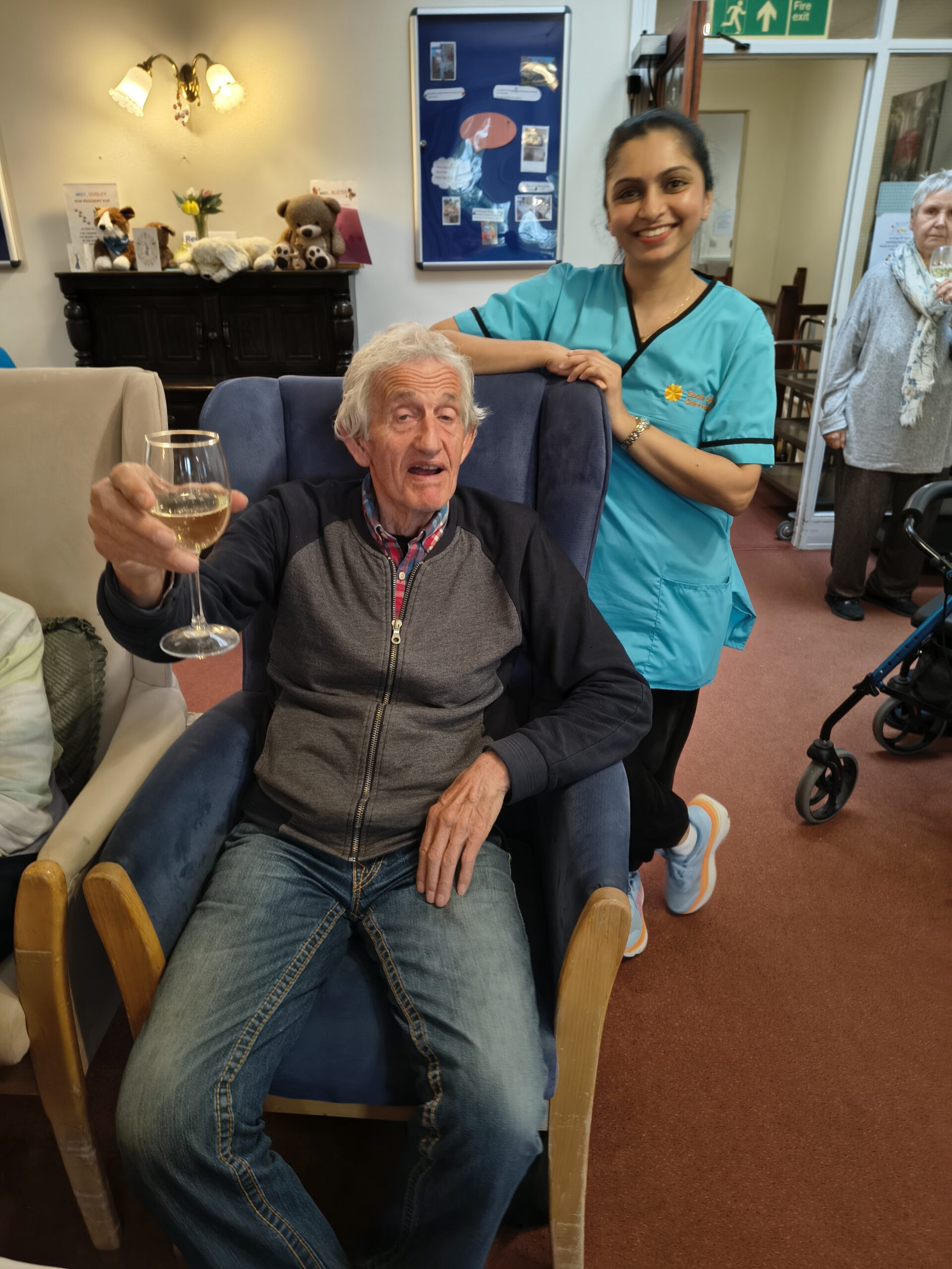 Male resident holding up a glass of bubbly with carer smiling Male resident holding up a glass of bubbly with carer smiling scaled