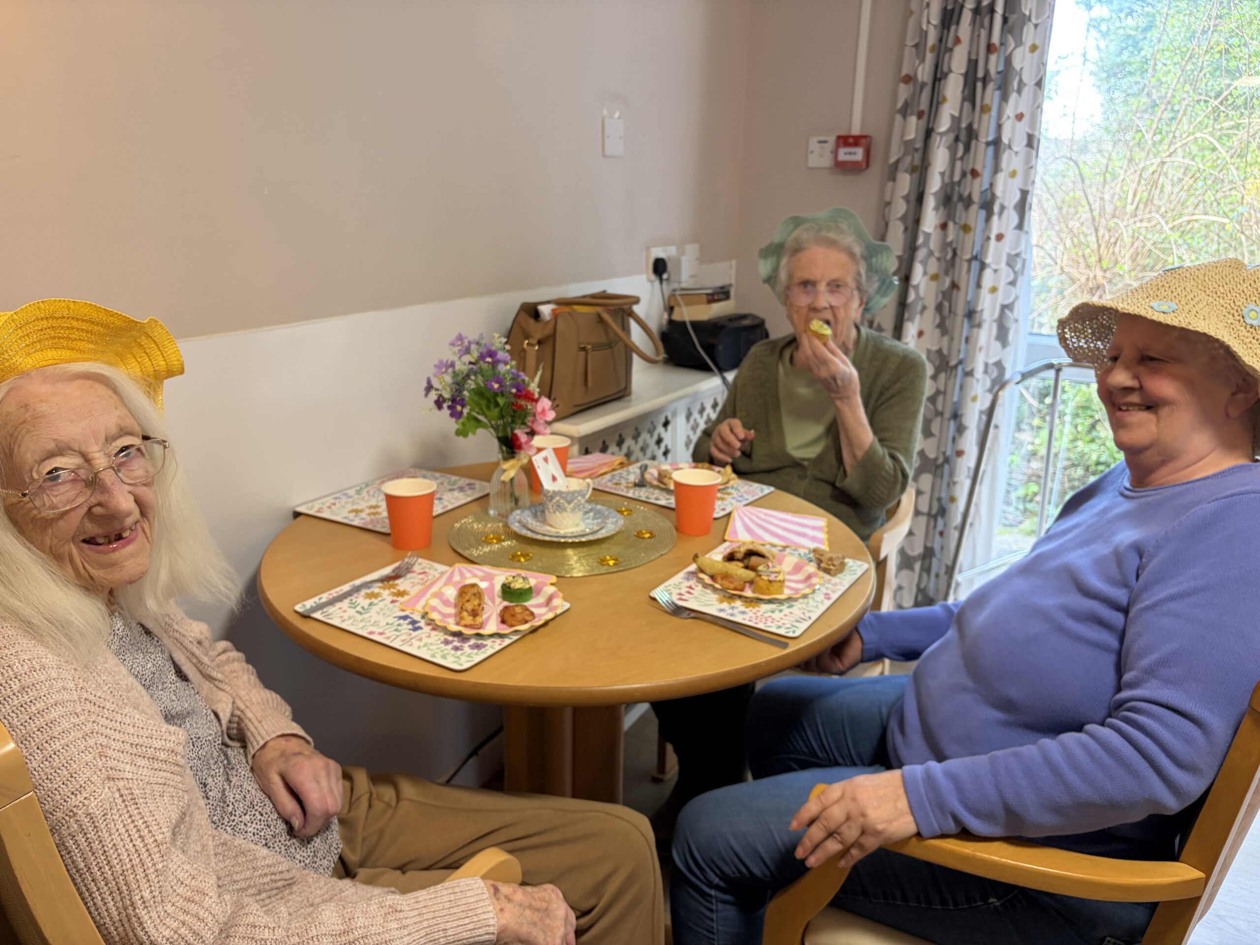 Three residents seated around a table enjoying the party food wearing hats scaled