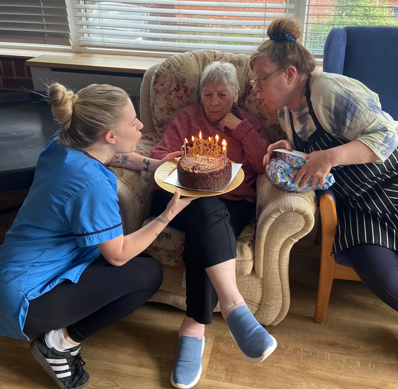 Ange seated with birthday cake blowing out the candles with help from Katie and one other