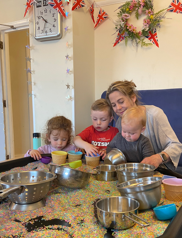 Claire and three children playing with a large tray of colourful rice and kitchen pans