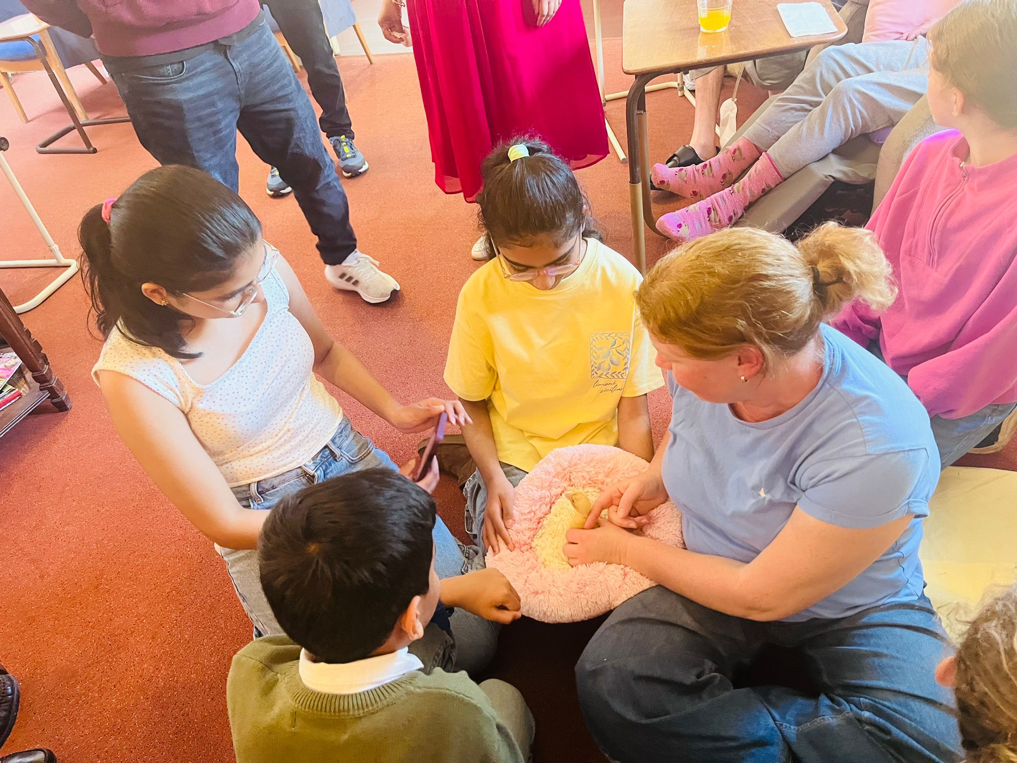 Group of children gathered around a carer holding a duckling on a pink fluffy pillow with residents watching