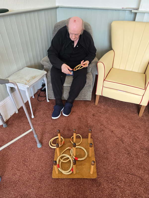 Maurice seated having a go throwing rope rings onto a board of wooden pegs on the floor