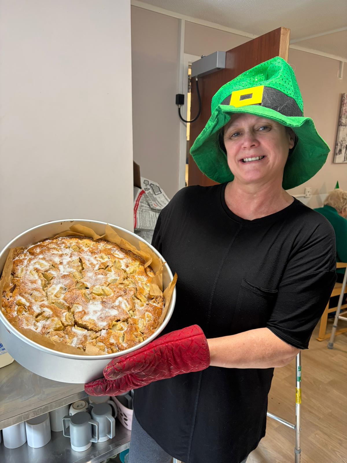 Michaelstowe staff member holding the apple cake using oven gloves wearing a leprechaun hat