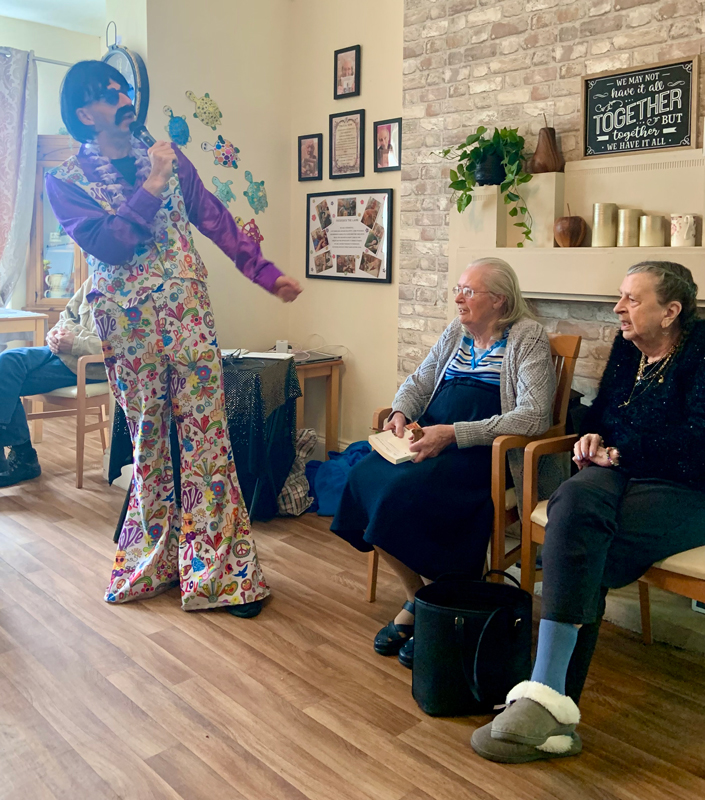 Paul dressed in a purple shirt and 60s print flares and waistcoat singing to residents