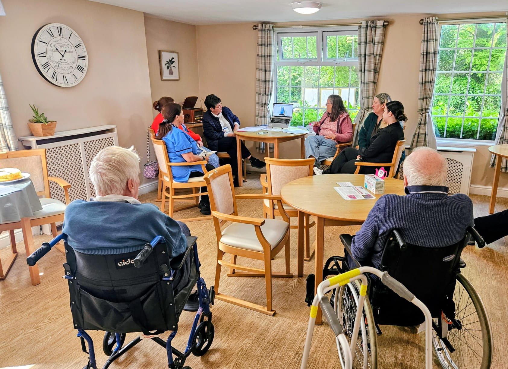 Residents and carers seated around tables of cakes watching a presentation on Dementia UK