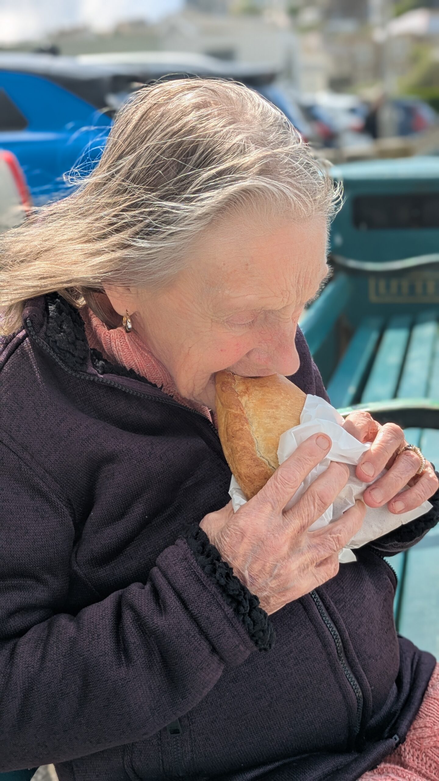 Thelma seated on a bench eating a Cornish pasty scaled