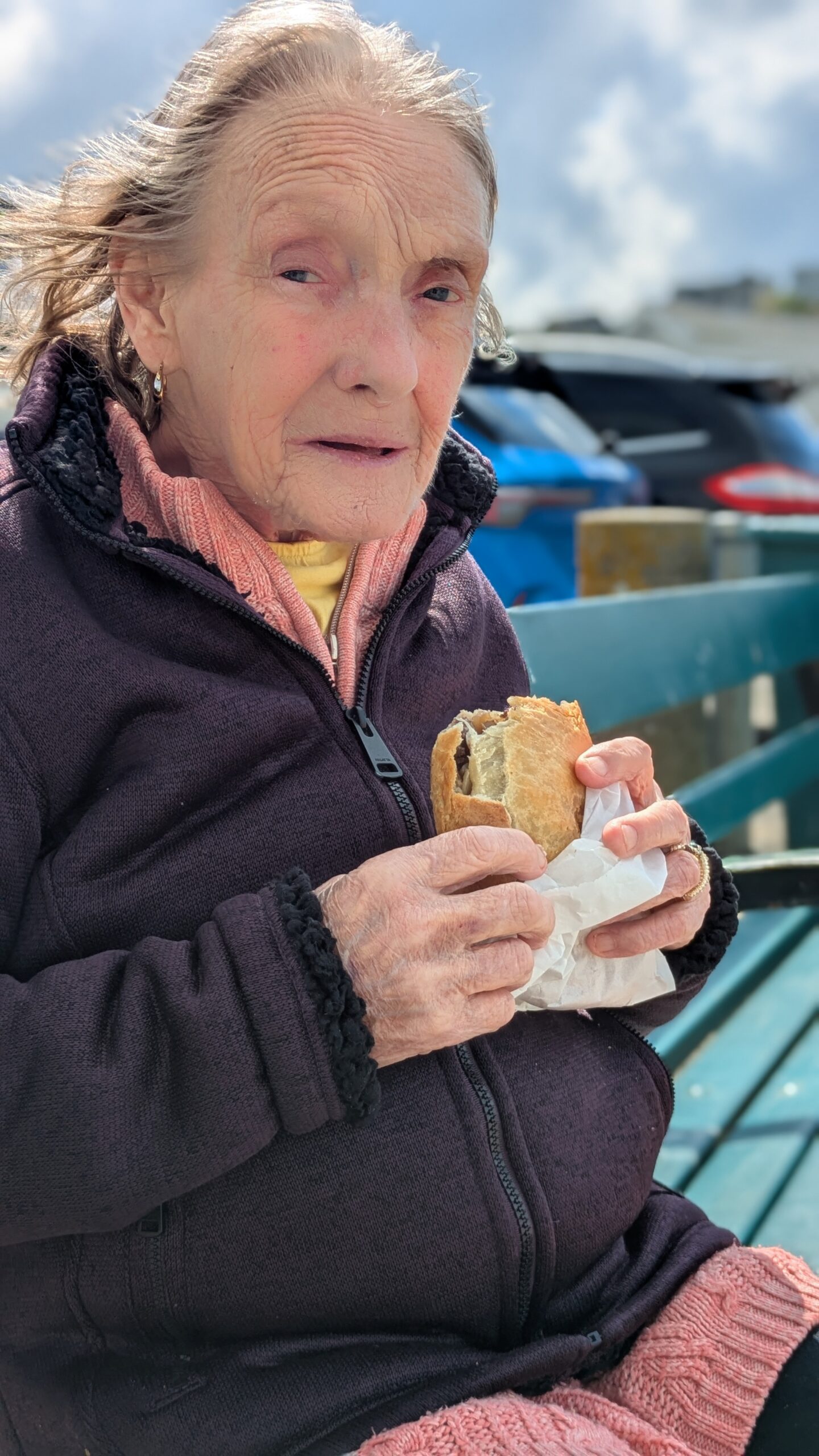 Thelma seated on a bench with a Cornish pasty enjoying the view scaled
