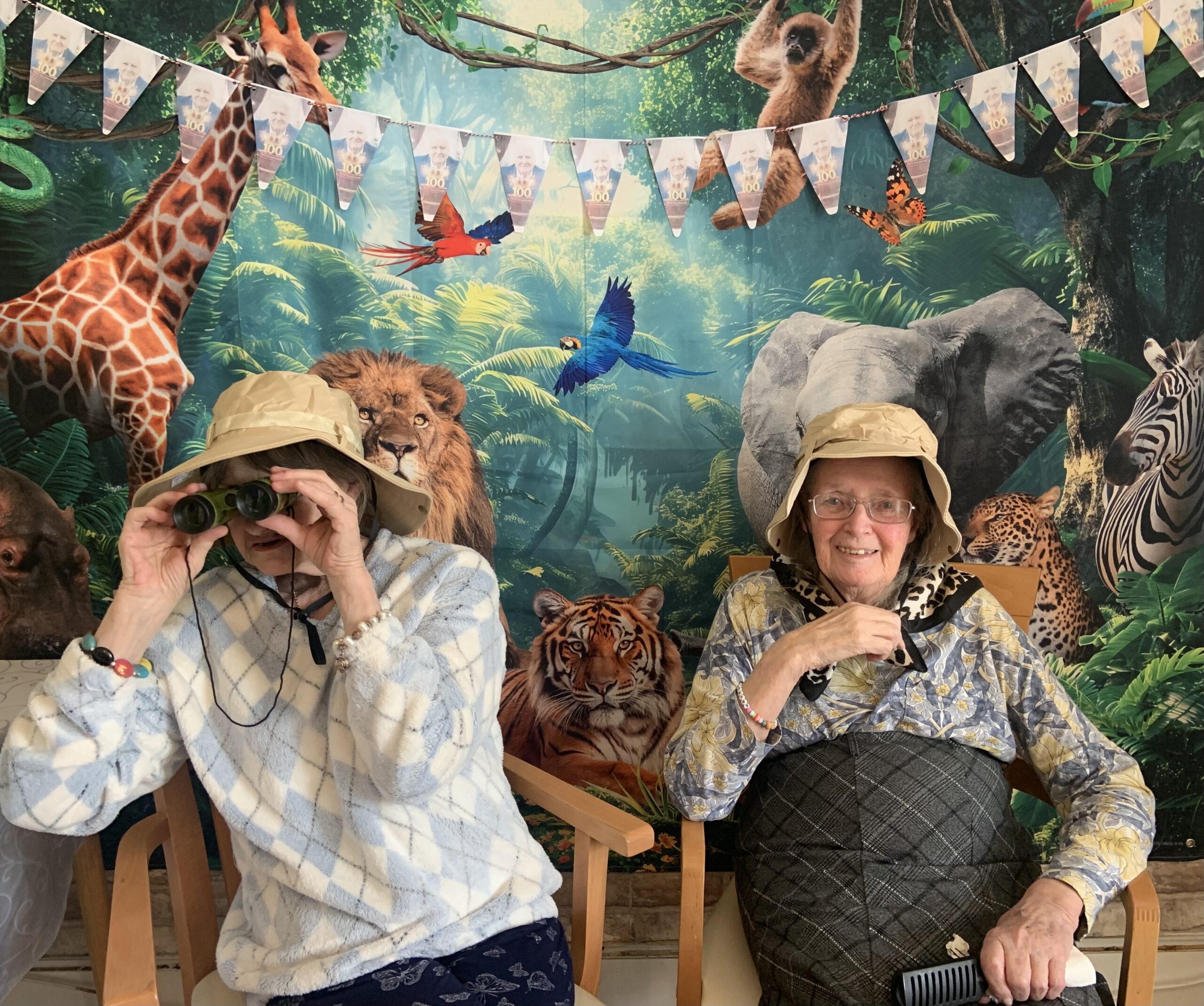 Two female residents in safari hats one with binoculars seated in front of jungle back drop scaled