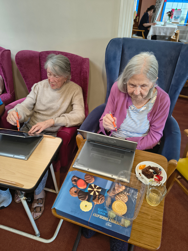 Two female residents seated in comfy armchairs with tables in front of them with tablets holding pens
