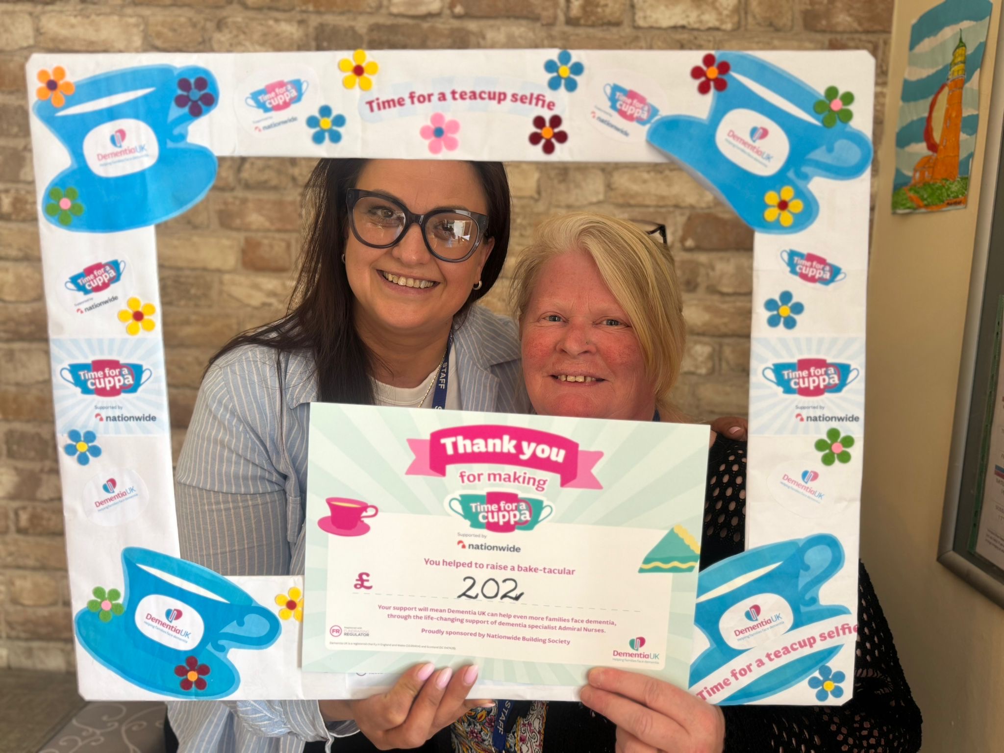 Two members of staff behind a Time for a Cuppa frame holding certificate thanking for donations