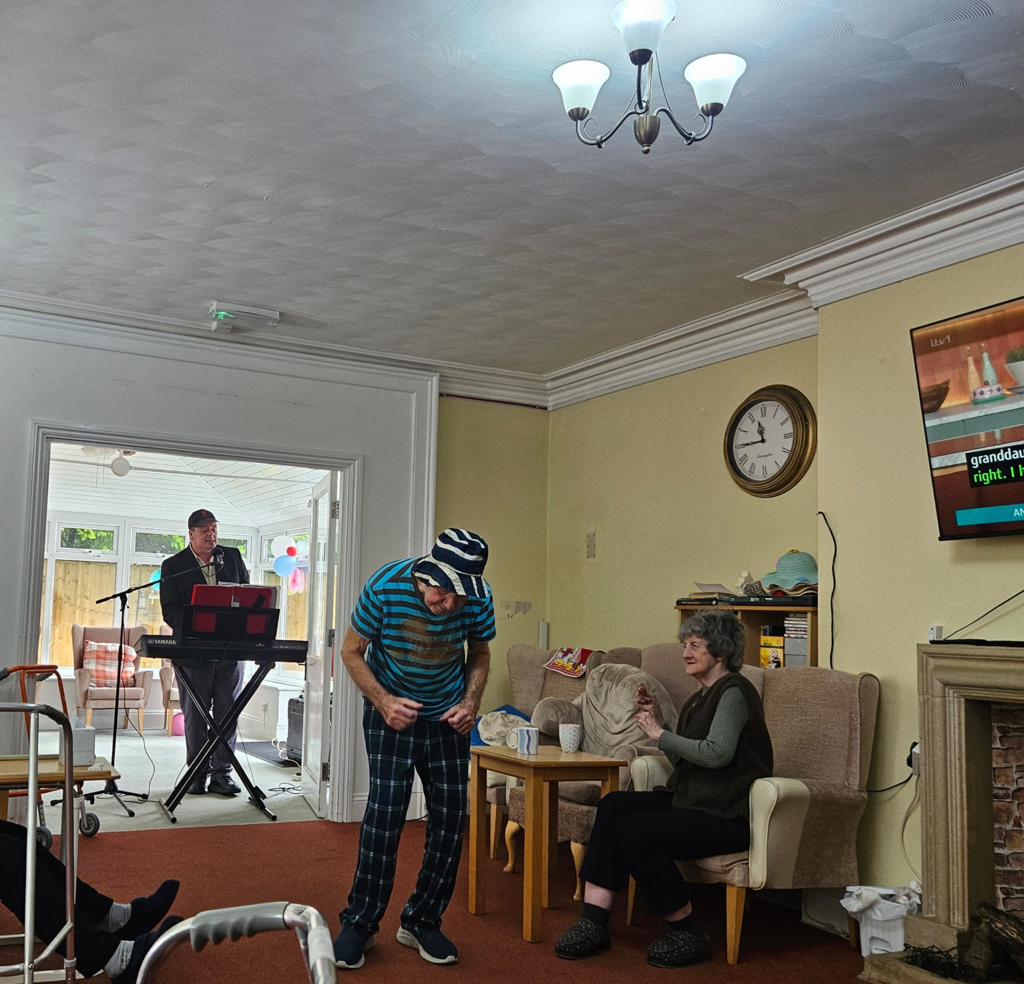 Wider shot of the same room with male resident dancing and one seated enjoying the music